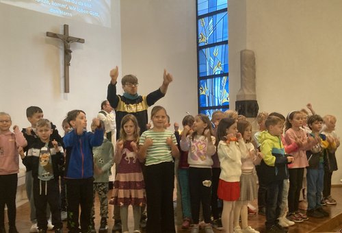 Klasse 2 singt mit ihrem Lehrer das Anfangslied. Sie untermalen das Lied mit Bewegungen. Im Hintergrund sind Kirchenfenster und Kreuz zu sehen.